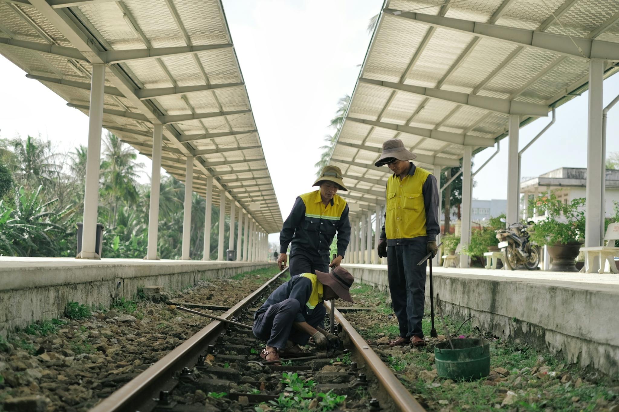 Three workers in uniforms maintaining railway tracks at a station under a canopy.